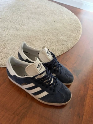 Adidas Gazelle blåvita sneakers - Säljer ett par klassiska Adidas Gazelle sneakers i blå mocka med vita detaljer och de ikoniska tre ränderna på sidan. Skorna har vit sula och plös samt mörkblå snören. Perfekta för dig som gillar retrostil och streetwear.