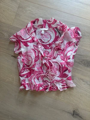 Blommig kortärmad blus från H&M Xs - Söt blus från H&M i transparent, lätt material med rosa och röda blommönster. Modellen har korta ärmar, volangdetaljer vid knäppningen och knytband i midjan. Perfekt för en trendig och somrig look.