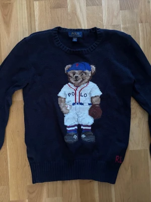 Mörkblå stickad tröja Polo Bear - Mörkblå stickad tröja från Polo Ralph Lauren med den ikoniska Polo Bear i basebolloutfit på bröstet. Tröjan har rund hals, ribbstickade muddar och RL-broderi nertill. (Barnstorlek)