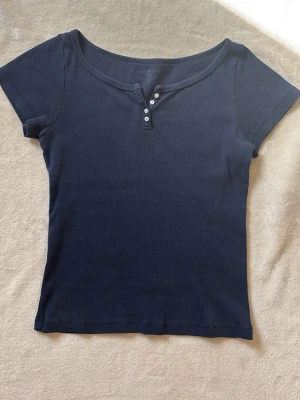 Mörkblå ribbad topp med knappar - Mörkblå ribbad topp från Brandy Melville med korta ärmar och rundad halsringning. Toppen har dekorativa knappar framtill och är tillverkad i mjuk bomullsmix. Har inte kommit till användning och är därför i mycket bra skick. Kunde inte hitta nypris men minns att den låg runt 280kr.🫶🏻