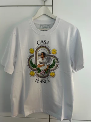 Vit t-shirt från Casablanca - Vit t-shirt från Casablanca med färgglatt tryck framtill som visar en tennisspelare, tennisbollar och gröna detaljer. Rund halsringning och korta ärmar. Materialet är mjukt och ser ut att vara bomull.