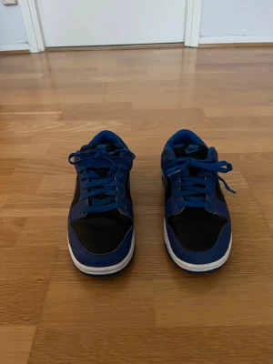 Nike Dunk Low Blå/Svart Sneakers - Storlek 41 - Nike Dunk Low blå/svart i storlek 41 säljes. Skorna är i bra skick med normala bruksspår från användning men har mycket kvar att ge. Den klassiska färgkombinationen gör dem lätta att matcha med olika outfits och passar lika bra till vardags som till fritiden. Bekväma, stilrena och redo för en ny ägare.