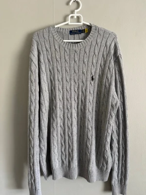 Ljusgrå tröja Polo Ralph Lauren - Säljer en ljusgrå kabelstickad tröja från Polo Ralph Lauren. Tröjan har rund halsringning, långa ärmar och den klassiska broderade loggan på bröstet. Tillverkad i mjukt material och har ribbade muddar vid ärmslut och nederkant. Har använts en fåtal gånger men är i riktigt fint skick. Storlek S