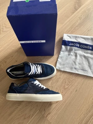 Jacob Cohën skor - Snygga mörkblå sneakers från Jacob Cohën med vit sula och vita skosnören. Ovandelen är i en mix av mocka och textil som ger en exklusiv känsla. Klassisk låg modell som passar till det mesta. Levereras med originalkartong och dustbag.