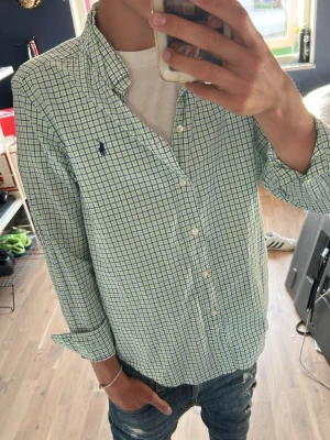 Grönrutig skjorta från Ralph Lauren - Säljer en klassisk grön- och vitrutig skjorta från Ralph Lauren med blå logga broderad på bröstet. Skjortan har lång ärm, knappar framtill och en avslappnad passform. Perfekt till jeans eller chinos för en clean look.