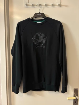 Svart sweatshirt från Adidas - Svart sweatshirt från Adidas med broderad logga och emblem på bröstet. Tröjan har rund halsringning, långa ärmar och ribbade muddar. Perfekt för en sportig och avslappnad stil.