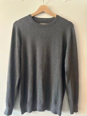 grå stickad tröja FMA Industries - Mörkgrå stickad tröja från FMA Industries med rund halsringning och ribbade muddar. Tröjan är enkel och stilren, perfekt för lager på lager och har en mjuk premiumkänsla. Passar till både jeans och chinos.