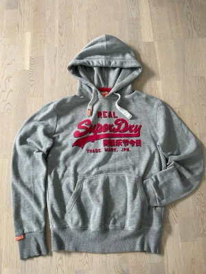 Grå Superdry hoodie med vinrött tryck - Snygg grå hoodie från Superdry med stort vinrött tryck på bröstet. Klassisk känguruficka framtill, vita dragsnören i huvan och ribbade muddar. Perfekt för en avslappnad och trendig stil. Materialet känns mjukt och bekvämt.