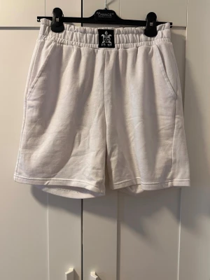 Vita mjukisshorts med resårmidja - Vita mjukisshorts med avslappnad passform och resår i midjan. Shortsen har två sidofickor och är tillverkade i ett mjukt bomullsmaterial. Perfekta för chill dagar eller när du vill ha det bekvämt.