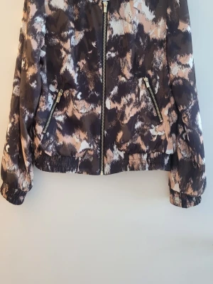 Mönstrad vindjacka med dragkedja från H&M. - Snygg vindjacka med abstrakt mönster i svart, beige och vitt. Jackan har dragkedja framtill, två fickor med dragkedjor och resår vid ärmslut och nederkant. Perfekt för dig som vill sticka ut med en unik look.