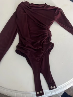 Vinröd långärmad body med drapering - Snygg vinröd body med långa ärmar och draperad detalj framtill. Tillverkad i ett mjukt, stretchigt tyg som ger en skön passform. Praktisk knäppning med tryckknappar i grenen. Perfekt för dig som vill ha en trendig och stilren look.