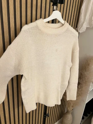 Krämvit oversized stickad tröja - Mysig oversized stickad tröja i krämvit färg med bred ribbad halsringning och muddar. Tröjan har en avslappnad passform och är perfekt för lager på lager. Materialet ser ut att vara mjukt och fluffigt, troligen en ull- eller akrylblandning.