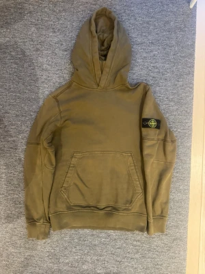 Olivgrön hoodie från Stone Island - Säljer en olivgrön hoodie från Stone Island med klassisk patch på vänster ärm. Hoodien har stor känguruficka framtill, ribbade muddar och en rejäl huva. Tillverkad i mjuk bomull för skön känsla och avslappnad stil. Storlek är 14 år men det blir som xxs.