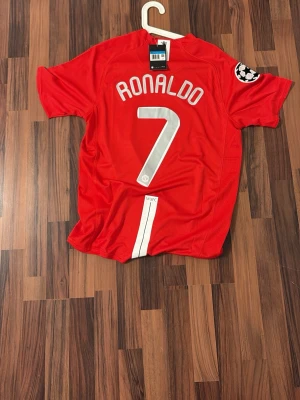 Manchester United Ronaldo 2007-8 - Röd Manchester United matchtröja med Ronaldo 7 på ryggen. Tröjan har vita detaljer, Champions League-märke på ärmen och AIG som sponsor på bröstet. Tillverkad i lätt och ventilerande material, perfekt för fotboll eller samling.