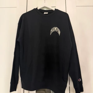 Svart sweatshirt från Champion - Svart sweatshirt från Champion med vit logga tryckt på bröstet och stor CHAMPION-text på ryggen. Köptes för 800kr . Klassisk rund halsringning och ribbade muddar vid ärmslut och nederkant. Perfekt för en avslappnad streetwear-look.