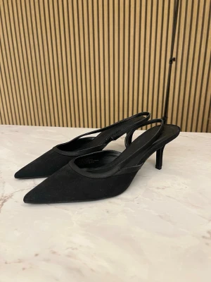 Svarta spetsiga slingback-pumps - Stilrena svarta pumps med spetsig tå och smal klack. Skorna har öppen häl med slingback-rem och är tillverkade i mockaimitation. Perfekta för dig som gillar en elegant och trendig look.