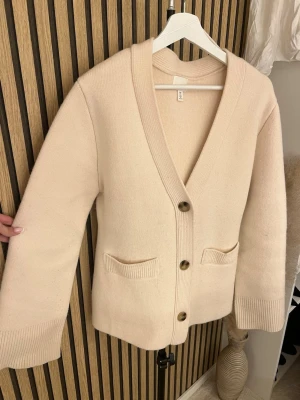 Beige stickad ull kofta med knappar - Snygg beige stickad kofta med v-ringning och stora knappar framtill. Två små fickor framtill och bred ribbad kant vid ärmslut och nederkant. Perfekt att slänga över en t-shirt eller topp för en chill look.