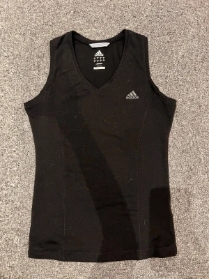 Svart träningslinne från Adidas - Snyggt svart träningslinne från Adidas med V-ringning och diskret logga på bröstet. Tillverkat i funktionsmaterialet Climalite som transporterar bort fukt, perfekt för gymmet eller löprundan. Ärmlös design för maximal rörelsefrihet.