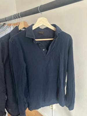 ROSE&BORN - Marinblå långärmad henley i bomull - Snygg marinblå långärmad henley med krage och knappar framtill. Tillverkad i bomull. Köpt från Rose&Born butik, knappt använd, kommer från tvätten därmed skrynklig. Stryks innnan frakt. **skriv för fler bilder** NYPRIS: 2400 kr