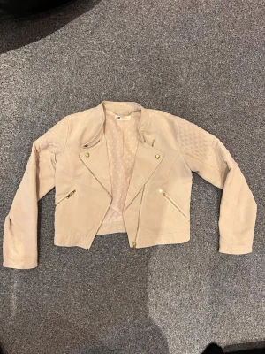 Beige skinnjacka från H&M - Snygg beige skinnjacka från H&M med guldfärgade dragkedjor och knappar. Jackan har quiltade detaljer på axlarna och ett kortare snitt. Fodret är ljust med små hjärtan. Perfekt för dig som vill ha en stilren och trendig look.