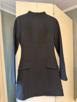 Svart klänning från Gina - Stilren svart kort klänning med lång ärm och hög hals. Klänningen har figurnära passform, dekorativa ficklock framtill och dragkedja i ryggen. Perfekt för dig som gillar minimalistisk och klassisk stil.