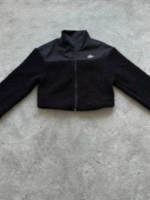 Svart teddyjacka från Alo - Säljer en svart teddyjacka från Alo med hög krage och dragkedja framtill. Jackan har en cool mix av mjukt teddy-material och slät panel upptill med Alo-logga på bröstet. Croppad modell som ger en trendig look och passar perfekt till lager på lager.