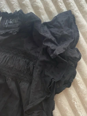 Svart blus med volang från Vero Moda - Snygg svart blus i 100% bomull från Vero Moda med volangdetaljer vid ärmar och hals. Spetsdetalj i fram. Blusen har prickigt mönster i tyget. Perfekt till sommaren, luftigt och skönt material. Använd en gång men aningen för liten så säljer den därför. Storlek 36! 