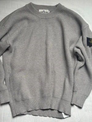 Grå stickad tröja från Stone Island - Snygg grå stickad tröja från Stone Island med ribbad struktur och rund hals. Tröjan har lång ärm och den ikoniska Stone Island-loggan på vänster ärm. Perfekt för dig som gillar stilrena och klassiska plagg.