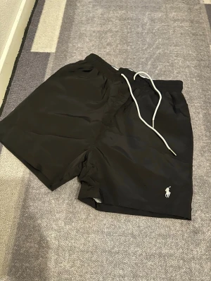 Svarta shorts från Polo Ralph Lauren - Svarta shorts från Polo Ralph Lauren med vit logga broderad framtill. Shortsen har elastisk midja med vit snörning och fickor på sidorna samt bak. Perfekta för sommaren och har en klassisk, enkel design.