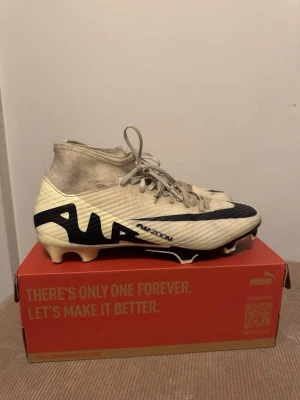Nike Zoom Mercurial fotbollsskor - Snygga Nike Zoom Mercurial fotbollsskor i beige och svart med hög ankel och snörning. Skorna har en modern design med stora svarta detaljer.