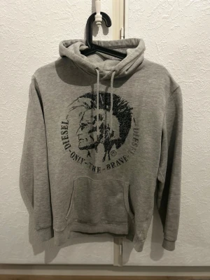 Grå hoodie från Diesel med tryck - Säljer en grå hoodie från Diesel med stort svart tryck framtill och texten 'Only The Brave'. Hoodien har huva med dragsko och en stor magficka. Perfekt för dig som gillar streetwear och vill ha något bekvämt och snyggt.
