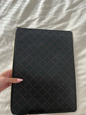 Svart datorfodral med rutmönster - Snyggt svart datorfodral med diskret rutmönster och guldfärgad dragkedja. Fodralet är tillverkat i ett tåligt material och har ett smidigt innerfack för extra förvaring. Perfekt för att skydda din laptop på språng.