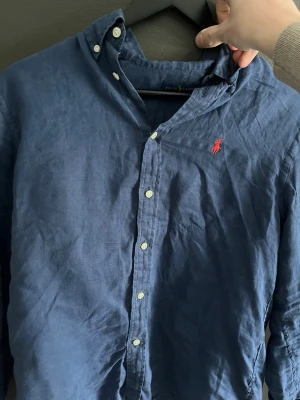 Blå linneskjorta från Ralph Lauren - Klassisk blå skjorta från Ralph Lauren i linne med button-down krage och vita knappar. Ikoniska röda Polo-loggan broderad på bröstet. Perfekt för en avslappnad men ändå stilren look.