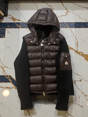 Marinblå Cardigan med huva från Moncler - Snygg marinblå Cardigan från Moncler med stickade svarta ärmar och huva. Västen har dragkedja framtill, ficka med Moncler-logga på ärmen och coola detaljer i metall. Perfekt för dig som vill ha en streetig och exklusiv look.