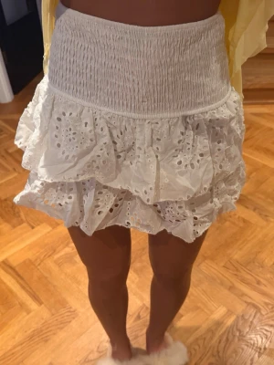 Vit volangkjol med broderi - Supersöt vit kortkjol med bred smock i midjan och dubbla volanger. Kjolen har broderade detaljer och hålmönster som ger en romantisk vibe. Perfekt för varma dagar och enkel att styla med en crop top eller skjorta.