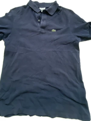Mörkblå pikétröja från Lacoste - Klassisk mörkblå pikétröja från Lacoste med korta ärmar och den ikoniska krokodilloggan på bröstet. Tröjan har krage och knappar framtill. Perfekt för en clean och sportig stil.