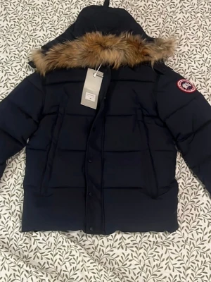 Mörkblå dunjacka från Canada Goose - Snygg mörkblå dunjacka från Canada Goose med avtagbar huva och pälsdetalj. Jackan har det klassiska Canada Goose-märket på ärmen och stora, praktiska fickor. Perfekt för kalla vinterdagar och riktigt varm tack vare dunfyllningen.