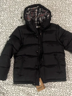 Svart pufferjacka från Burberry - Säljer en svart pufferjacka från Burberry med rutigt foder i grått, vitt och rött. Jackan har en stor huva med fuskpäls och dragkedja framtill. Märkeslogga på ärmen och praktiska fickor. Perfekt för kalla vinterdagar.