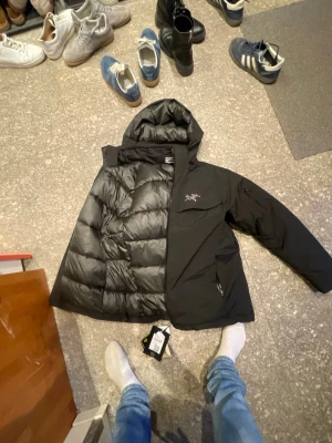 Svart dunjacka från Arc'teryx, Acteryx jacka - Snygg svart dunjacka från Arc'teryx med huva och dragkedja framtill. Jackan har en diskret logga på bröstet och är fodrad med dun för extra värme. Utrustad med flera fickor och tillverkad i slitstarkt material med GORE-TEX för väderskydd. Super nu iför skidor och kyla. Bara att höra av dig vid funderingar 