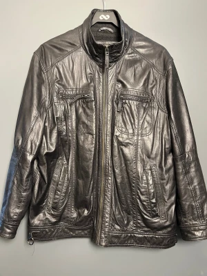 Svart skinnjacka från Levinsky - Snygg svart skinnjacka från Levinsky med hög krage och dragkedja framtill. Jackan har flera fickor med dragkedja både på bröstet och sidorna, samt detaljerade sömmar för en cool look. Perfekt för dig som vill ha en klassisk och stilren skinnjacka.