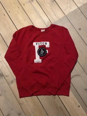 2000s Abercrombie & fitch hoodie - Snygg röd crewneck hoodie från Abercrombie & Fitch med stor vit broderad bokstav och svart patch med A&F-logga på bröstet. Tillverkad i mjukt bomullsmaterial och har ribbade muddar vid ärmslut och nederkant. Perfekt för en avslappnad och sportig look.