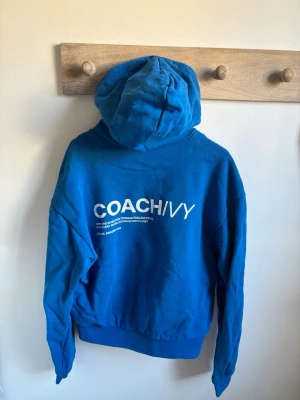 Blå hoodie från Coach Ivy - Snygg blå hoodie från Coach Ivy med vit logga på bröstet och större tryck på ryggen. Klassisk känguruficka framtill och stor huva. Tillverkad i mjukt bomullsmaterial, perfekt för en chill och avslappnad stil.