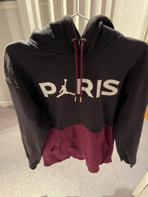 Jordan PSG hoodie svart vinröd - Snygg hoodie från Jordan x PSG i svart och vinrött med stor PARIS-text och Jumpman-logga på bröstet. Har huva med vinröda snören och en stor magficka. PSG-märke på ärmen. Tillverkad i mjuk bomullsblandning, perfekt för en sportig och trendig look.