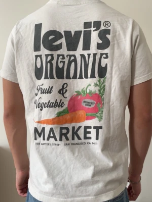 Vit Levi's t-shirt med tryck - Säljer en vit t-shirt från Levi's med stort färgglatt tryck på ryggen som föreställer texten 'Levi's Organic Fruit & Vegetable Market' och illustrationer av grönsaker. T-shirten har klassisk passform och är kortärmad, perfekt för en chill och trendig look. Storlek S