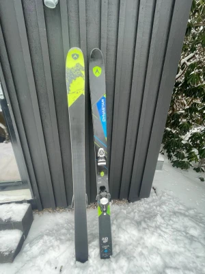 Dynastar Cham 97 allmountain skidor - Ett par Dynastar Cham 97 allmountain skidor med svart, blå och neongul design. Skidorna har bindningar monterade och är märkta med 'idecore' och 'CHAM 97'. Perfekta för dig som vill ha mångsidiga skidor till både pist och offpist.