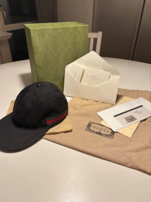 Svart keps från Gucci med monogram - Svart keps från Gucci med diskret GG-monogram över hela tyget och klassiska gröna och röda ränder på sidan. Kepsen har böjd skärm och justerbar rem baktill. Kommer med originalförpackning och dustbag.