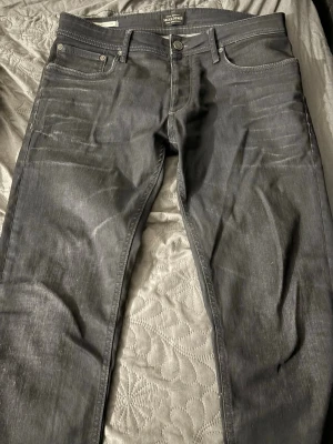Mörkgrå jeans från Jack & Jones - Snygga mörkgrå jeans från Jack & Jones med klassisk femficksmodell och raka ben. Jeansen har silverfärgade nitar och knapp, samt tydliga sömmar. Tillverkade i slitstarkt denimtyg som passar perfekt till en avslappnad stil.