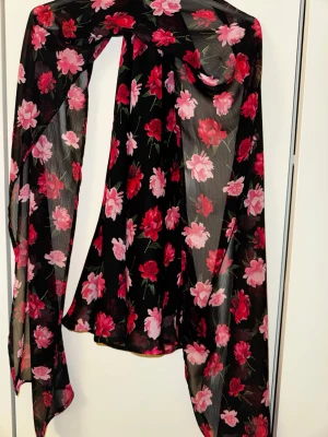 Svart blommig sjal i chiffong - Snygg svart sjal i tunn chiffong med blommigt mönster i rosa och rött. Perfekt att slänga över axlarna för en färgklick till outfiten. Lätt och luftig, passar till många olika stilar.