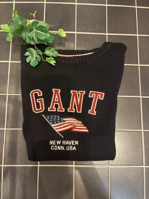 Svart stickad tröja från GANT - Snygg svart stickad tröja från GANT med broderad amerikansk flagga och texten 'NEW HAVEN CONN. USA' på bröstet. Tröjan har rund halsringning, långa ärmar och ribbade muddar. Perfekt för dig som gillar klassisk college-stil.