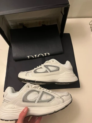Vita Dior  sneakers - Säljer ett par vita Dior  sneakers med gråa och silvriga detaljer. Skorna har en chunky sula, mesh och läderpaneler samt tydlig Dior-logga på sidan. Snygg och modern design som sticker ut. Perfekta för dig som gillar exklusiva sneakers och streetwear.